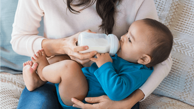 A mom in a beige sweater formula feeding her baby in a blue onesie