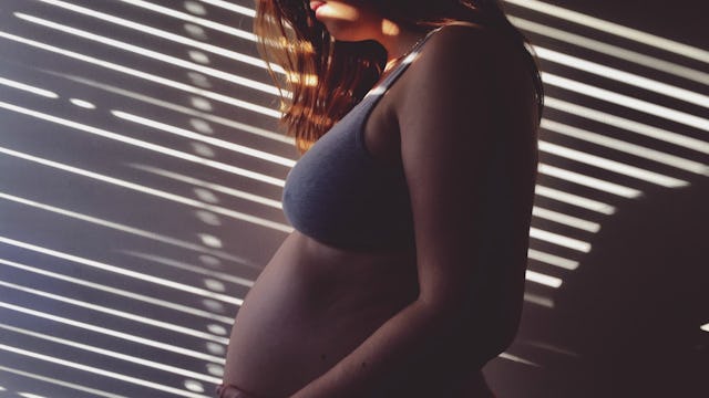 A pregnant woman standing alone in a shadow of blinds from a window