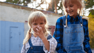 Two blonde young girls wearing shirts and denim overalls smiling while the younger one is picking he...