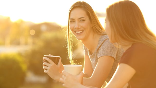 Two women leaning on a balcony during sunrise, with cups of coffee in their hands, talking and smili...