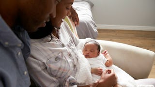 A dad sitting and hugging a mom on a white couch who is holding a newborn baby