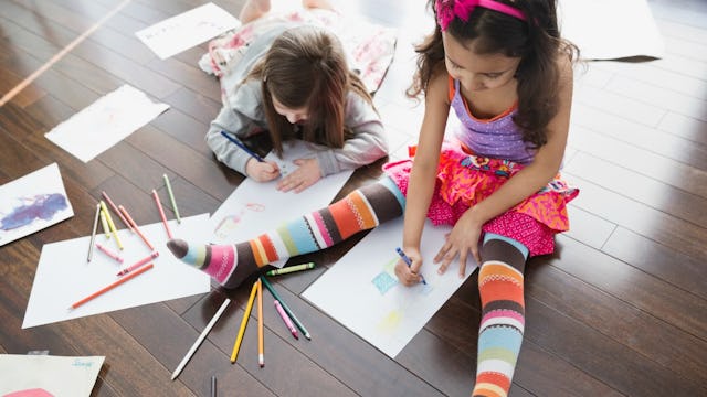 Two girls on a playdate, sitting and lying on the wooden floor while drawing something on a paper