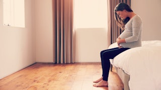 Pregnant woman in a grey shirt and black trousers that lost her closest friend sitting on a bed