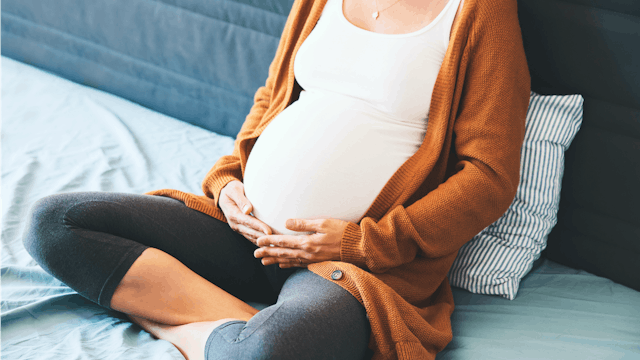 A pregnant woman in a white shirt, blue jeans, and orange sweater sitting and holding her belly as ...