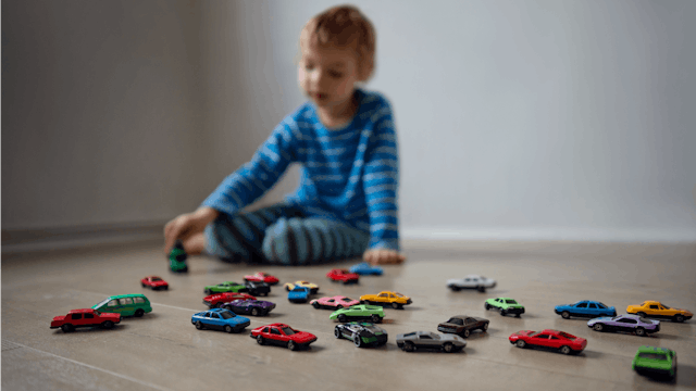 A child who doesn't speak in a blue shirt sitting on a wooden floor and playing with his toy cars