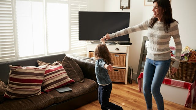 Mom, wearing a grey and white sweater and jeans, and daughter dancing joyfully in the living room.