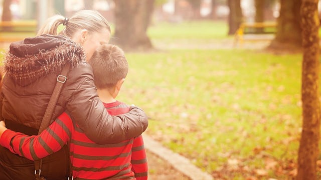 A woman in a brown puffer jacket hugging her son in a red and black striped sweater in a park