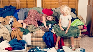 A child sitting and another one lying on a messy couch with lots of clothes thrown at the couch