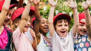 A group of children wearing red caps, laughing with their hands in the air at the daycare.