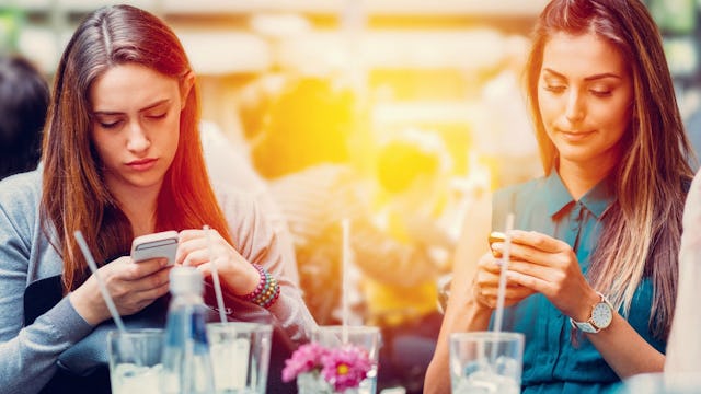 Two fake friends sitting with cocktails in front of them and both scrolling through their phones