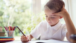 A blonde little boy in a white shirt sitting at a desk and doing his homework