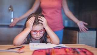 A young girl studying and holding her head in her hands; her mom is behind her with her arms open.,