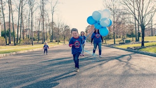 A boy with autism running holding a few blue balloons and mother and two of his sisters standing beh...