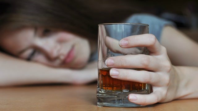 An alcoholic mom leaning her head on a table while holding a glass with liquor in it