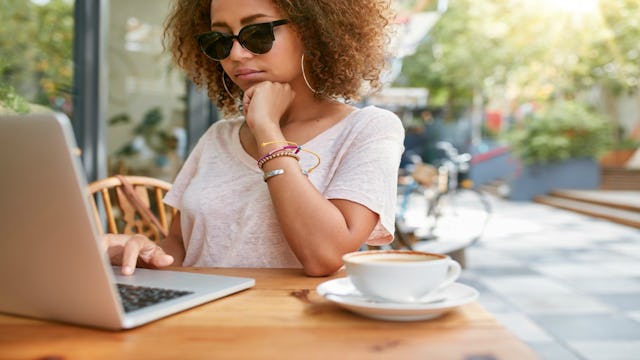 A girl with an Afro hairstyle wearing sunglasses sitting in a café drinking coffee while browsing so...