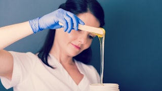 A black-haired beautician in a white shirt and blue gloves preparing wax for a treatment