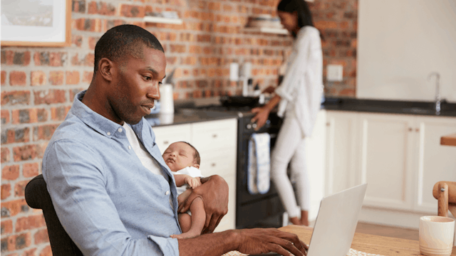 A working dad holding his baby in his arms and working on the laptop at the same time during the mot...