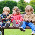 Brown haired mother wearing jeans watching her three children sitting on a bench while they are play...