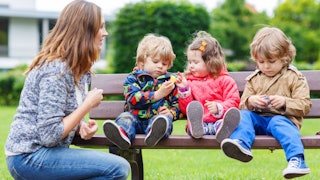 A mother wearing jeans watching her three children sitting on a bench while they are playing