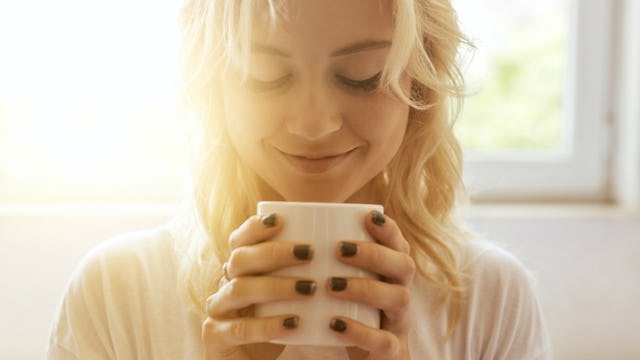 A blonde woman smiling and holding a cup, looking at it and smiling.