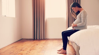 A single and pregnant woman sitting alone on the side of her bed with white sheets 