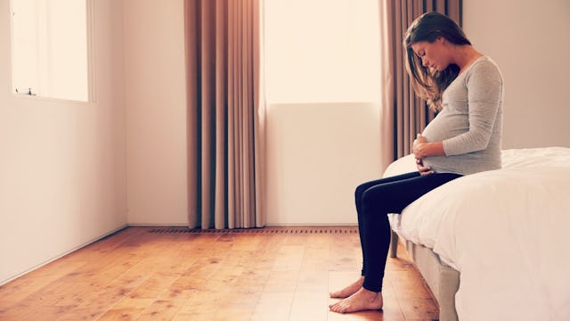 A single and pregnant woman sitting alone on the side of her bed with white sheets