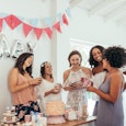 Five female friends standing next to a baby shower party sign and cake