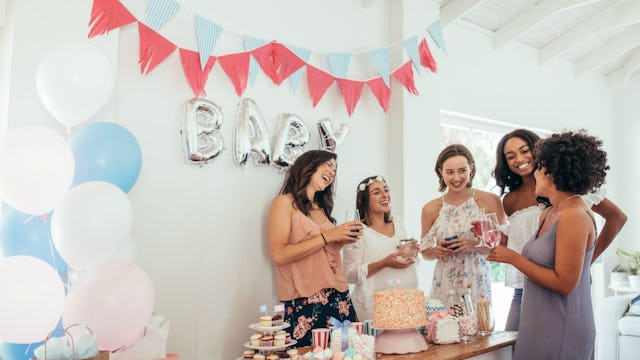 Five female friends standing next to a baby shower party sign and cake during a baby shower gatherin...