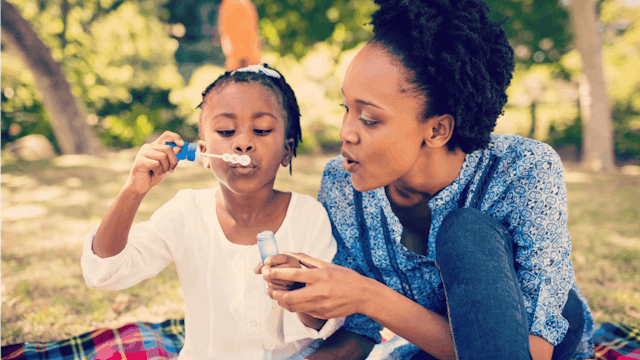 A childless stepmom sitting next to her stepdaughter, who is blowing bubbles in a park during a sunn...