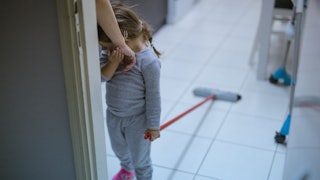 A toddler in a grey sweatshirt and pants holding a cleaning mop while leaning her face on her mother...