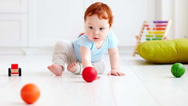 A red-haired toddler in a blue shirt and gray sweatpants playing on a floor with three balls around ...