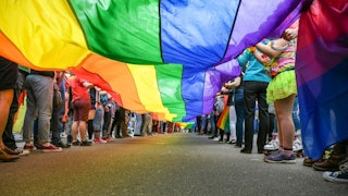 Kids standing in two lines while holding an oversized LGBTQ flag on both sides