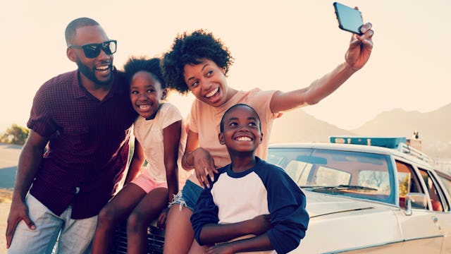 A man, woman, girl, and boy taking a family selfie on top of their car for a social media post