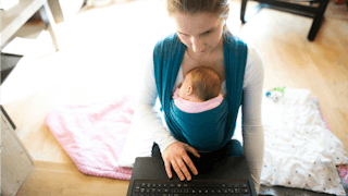 A mom carrying her baby in a teal baby wrap while sitting and working on her laptop