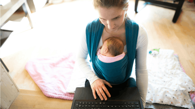 A mom carrying her baby in a teal baby wrap while sitting and working on her laptop
