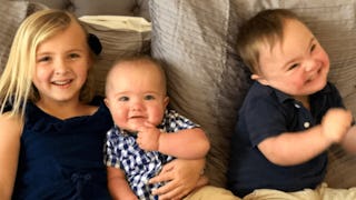 A blonde girl in a black top lying down holding a baby next to the middle child with Down Syndrome w...