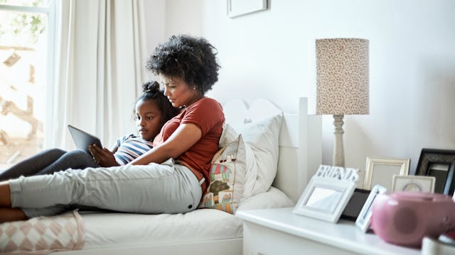 A mother lying on a bed with her daughter while they are watching something on a tablet in a bedroom...