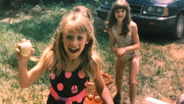 Two young girls happily playing in a sunny garden next to a parked gray car during Summer in the 90s...