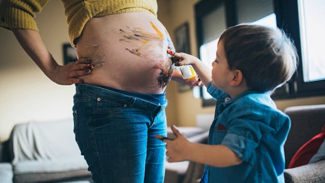 A pregnant woman holding some paint while her son paints on her exposed belly