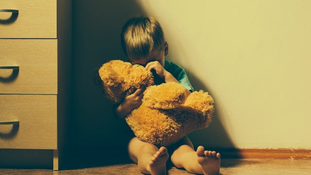 A boy who is anxious sitting in a corner of his room holding a large teddy bear