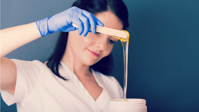 A brunette woman in a white T-shirt wearing blue gloves is preparing wax for a Brazilian Wax procedu...