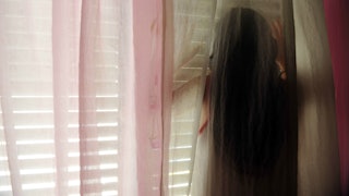 A tween girl behind the sheer curtains looking through the gaps in window shutters