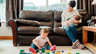 A mom breastfeeding a baby while her toddler plays on the floor next to them