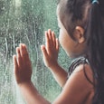 A small girl holding her hands leaning on a window who is masking anxiety with curiosity