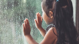 A curious child with pigtails, leaning her hands on a window and looking through during a rainy day