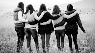 5 women from the back standing next to each other side to side, holding each other's backs and shoul...