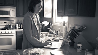 A black and white photograph of a mom looking at her laptop in the kitchen, feeling like a 'mom mart...