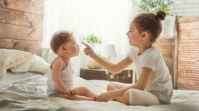 A toddler and a baby sister, who have a strong sibling bond playing with each other on a white bed