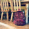 Black backpack with a pink outline and shapes leaned on a wooden chair at the table
