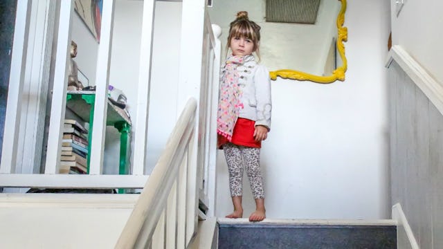 A sad child on top of white stairs in a white jacket, red shirt, leopard print tights and a geometri...